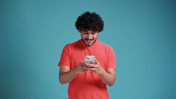 istock Dreamful marvelous fun young bearded Indian man 20s years old hold in hand use mobile cell phone typing browsing chatting send sms in coral t-shirt on blue studio background studio portrait 1483826033