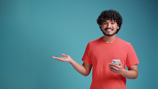 Young indian man using smartphone standing very happy pointing with hand and finger to the side, in coral t-shirt on blue studio background.