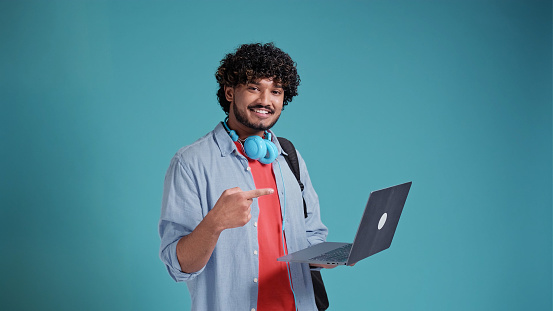 Handsome Indian man student on a blue background, holding a laptop with and pointing at him with a hand gesture