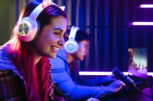 Woman smiling happily as she takes part in video gaming with a male player. Female gamer wearing a headset and sitting in front a microphone, live recording the exciting gameplay on an online streaming platform.