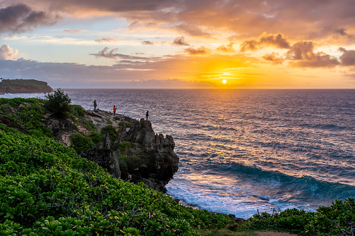 Men, fishermen, standing, fishing, on the edge of a cliff over the ocean, with the sun rising over the horizon and a partly cloudy sky, Mahaulepu Heritage Trail, Kauai, Hawaii