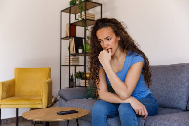 Anxious young woman sitting on the sofa, biting her nails, contemplating stock photo