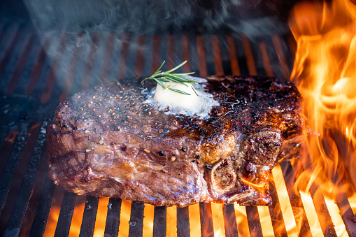Juicy rib eye beef steak cooked to perfection on a flaming grill with a dollop of butter and a sprinkle of rosemary. A succulent and flavorful meal