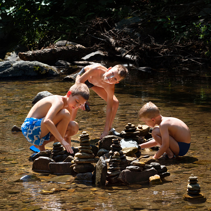Children happy among nature playing in water of river in summer hot day, building stone pyramids of pebbles. Summer holiday, vacation, free time activity outdor, fun and joy