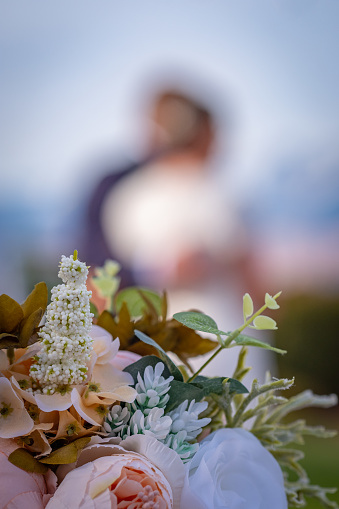 An iconic look at a couple sharing their first dance at their wedding. The shallow depth of field makes the couple out of focus and unrecognizable and is perfect for overlaying copy and/or logos. The wedding is outdoors so there are trees and blue sky in background. In focus in the foreground is a fresh bouquet of white and pink roses.