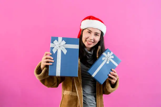 portrait, Asian woman wearing long-sleeved outerwear and Christmas hat, holding two gift boxes with two hands held out happily receiving two boxes presents, Isolated indoor studio on pinkbackground.