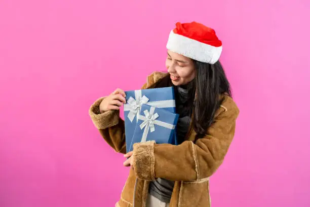 portrait, Asian woman wearing long-sleeved outerwear and Christmas hat, wrapped arms around two gift boxes on chest looked down with delight at receiving it, Isolated indoor studio on pinkbackground.