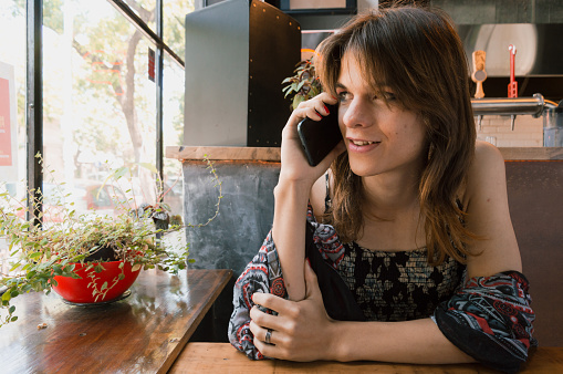 beautiful young transgender hispanic latina woman of argentinian ethnicity, in a restaurant sitting talking on the phone, sitting leaning on the table, calm, alone and looking out the window.