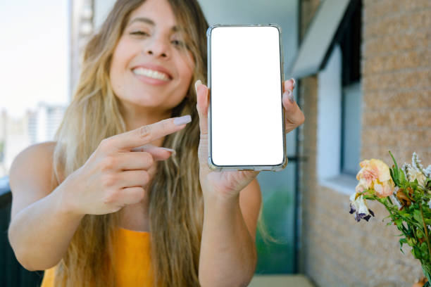 young woman smiling holding her phone pointing the blank screen with her finger. focus on the phone stock photo