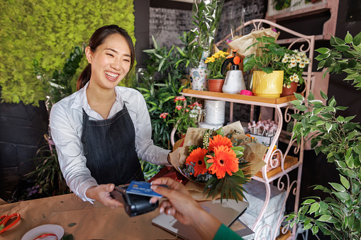 Smiling and friendly florist holding card reader machine at counter with customer paying with credit card