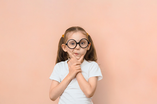 Cute funny little caucasian girl in round glasses and hands near her face is thinking something while looking at the camera on a pink background with copy space.
