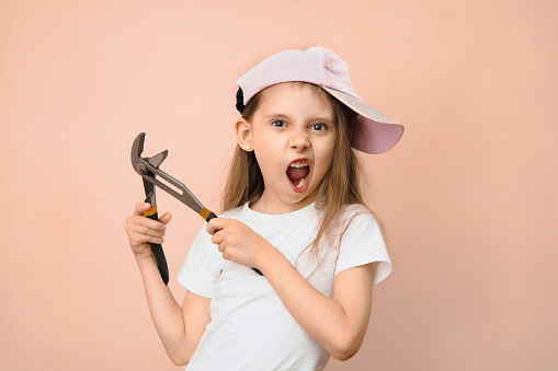 Caucasian girl of primary school age in a baseball cap with a wrench on a pink background, father's daughter tomboy.