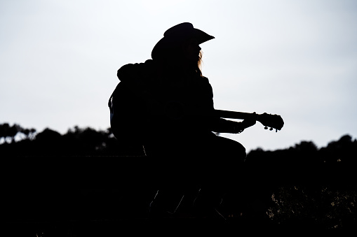 Silhouetted Woman Playing Guitar Outside Sitting on Fence - Playing guitar outdoors backlit against bright background.