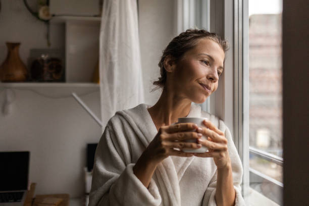 istock Young woman at home drinking coffee wearing robe looking through window 1483916000