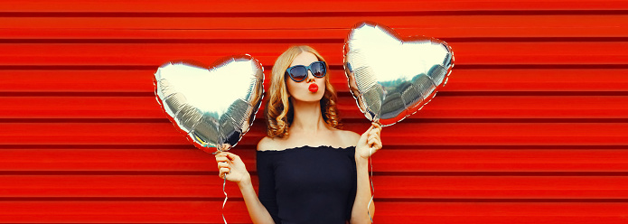 Portrait of beautiful young woman blowing her lips sending sweet air kiss with silver heart shaped balloons in hands on red background