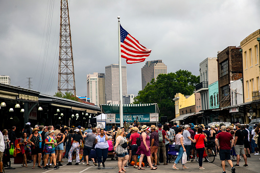 New Orleans April 15,2023:  New Orlena's French Quarter Farmers Market.  Crowd outside the market with large American Flag overhead.    Grey overcast sky/