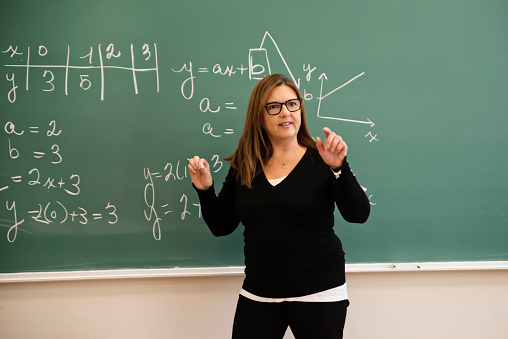 Senior female math teacher writing on board in classroom. She is in her fifties. This is part of a series promoting diversity and equity in STEM. Horizontal waist up indoors shot with copy space. This was taken in Montreal, Quebec, Canada.