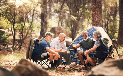 Man, friends and camping in nature laughing for funny joke, meme or conversation by tent in forest. Group of elderly men relaxing on camp chairs with drink enjoying sunny day together in the woods