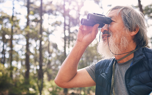 Binoculars, senior man and hiking in nature looking at view, sightseeing or watching. Binocular, adventure search and elderly male with field glasses, trekking or exploring on vacation outdoors.