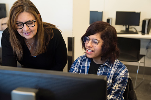 College student coding with teacher in technology lab classroom. Student is a transgender young woman and teacher is a senior woman. This is part of a series promoting diversity and equity in STEM. Horizontal waist up indoors shot with copy space. This was taken in Montreal, Quebec, Canada.