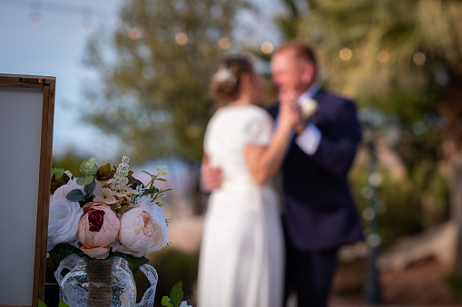 An iconic look at a couple sharing their first dance at their wedding. The shallow depth of field makes the couple out of focus and unrecognizable and is perfect for overlaying copy and/or logos. The wedding is outdoors so there are trees and blue sky in background. In focus in the foreground is a fresh bouquet of white and pink roses.
