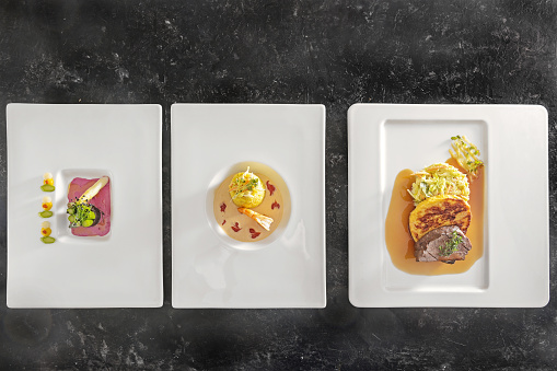 Three menu dishes on white plates, asparagus with beetroot sauce, saffron risotto with prawn, roast deer with dumpling and vegetable, dark background, top view from above, copy space, selected focus