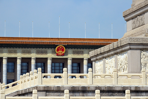 Beijing, China: Great Hall of the People façade with Chinese coat of arms, seen behind the Monument to the People's Heroes. One of the most symbolic buildings in Beijing. The neoclassical-style building designed by Zhang Bo is located on the west side of Tiananmen Square and serves the Chinese leadership as a place to receive state guests and as a venue for national celebrations and as the meeting place of the Chinese parliament (National People's Congress).