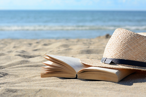 Open book and straw hat on sandy beach near sea, space for text