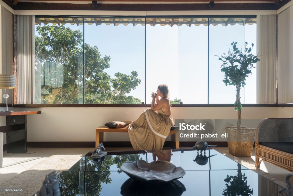 Mature woman drinking by hotel window Mature woman drinking coffee by a large window. In a hotel room with view to trees Fashion Stock Photo