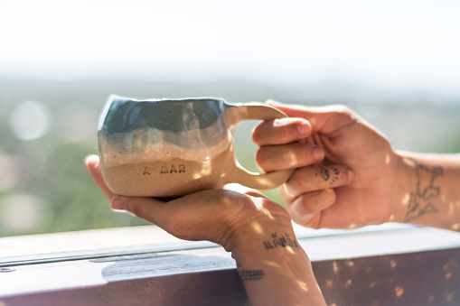 holding a coffee cup in the morning, by a window. Tattoed hands