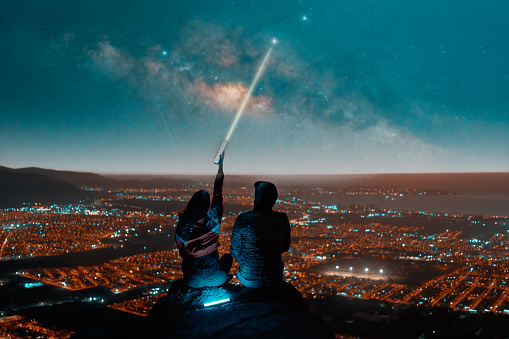 silhouette of heterosexual couple sitting on top of mountain watching pointing out with laser the starry sky with milky way over the city