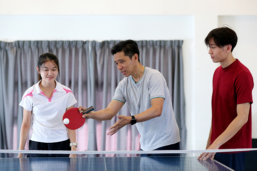 An Asian man is coaching young man and young woman skills on table tennis (ping pong)