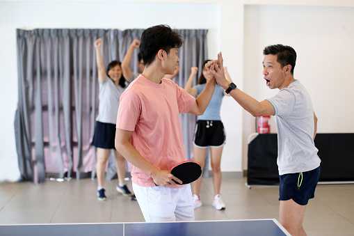 An Asian man is celebrating winning with his teammate playing table tennis (ping pong) while fans cheering at background