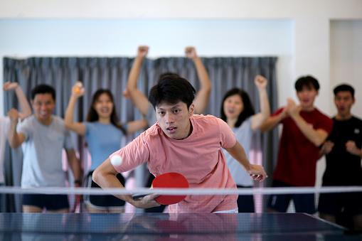 Asian young adult is enjoying playing table tennis with spectators cheering at background
