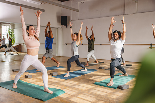 An adult caucasian female yoga practitioner showing her diverse clients a yoga pose during their morning yoga class. They are performing a warrior yoga pose and are therefore standing on yoga mats in a lunge position with their arms extended over their head. The class is taking place in a cute yoga studio with a big mirror and big windows.