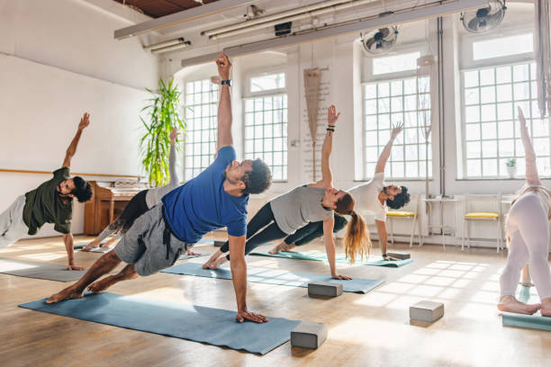 diversos participantes de la clase de yoga haciendo una tabla lateral en sus colchonetas de yoga en un hermoso estudio de yoga con grandes ventanales - pilates fotografías e imágenes de stock