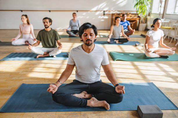 diversi partecipanti alla lezione di yoga che praticano la posa di sukhasana in un piccolo studio di yoga accogliente con luce naturale - yoga class instructor yoga exercising foto e immagini stock