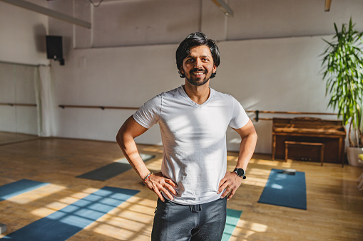 Happy Indian male yoga instructor standing with his hands on his hips and smiling at the camera. He is located in a cosy studio with natural light.