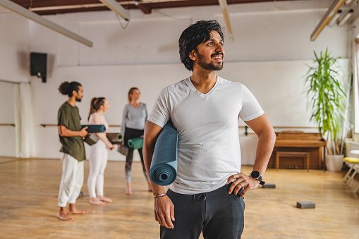 A portrait of an attractive male yoga instructor looking away from the camera. He is located in a beautiful simple yoga studio with big windows. In the background there are some  of his yoga clients having a discussion.