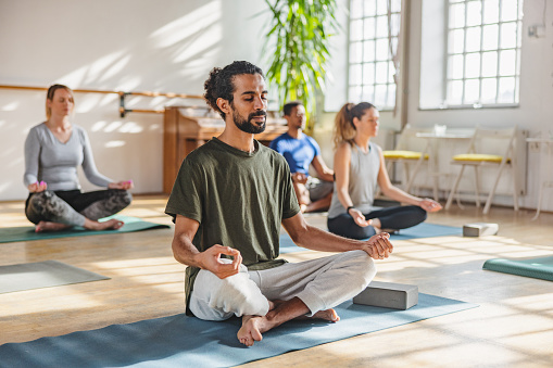 An adult arab male with a ponytail meditating in a yoga class. He is surrounded by othe yoga class particpants that are diverse in age, gender and race. The yoga practitioner looks calm and relaxed. The class in taking place in a cosy bright studio with big windows and plants.