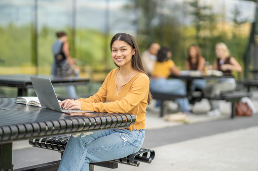 A young female University student, sits at a table outside as she studies between classes.   She is dressed casually and smiling.