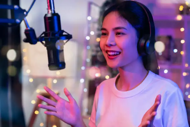 Photo of Cheerful beautiful Asian woman wearing headphones and sing through the microphone with using laptop record podcast. Content creator concept.