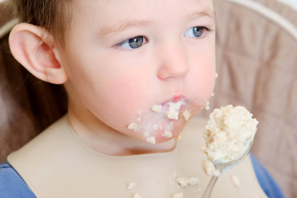 istock A happy child eats porridge with a spoon while sitting on a high chair. Baby in a bib eats oatmeal porridge himself. Kid aged about two years (age one year nine months) 1484020861