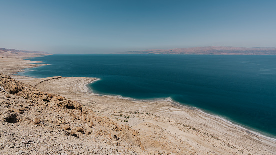 Israel Dead Sea Coastal Panorama. View from above along the dry rocky Israel Dead Sea Coastline of the West Bank in Israel. Jordan Dead Sea Coast in the Background. Dry rocky desert coast and deep blue salty water of the Dead Sea under blue cloudless summer skyscape. Dead Sea, Mineral Beach, Ein Gedi, Israel, Middle East