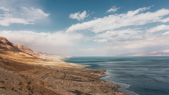 Israel West Bank Dead Sea Coast Panorama. Scenic view from above along the dry rocky Israel Dead Sea Coast in Israel. Dry rocky desert mountain range and coast towards the deep blue salty water of the Dead Sea under blue skyscape with fluffy clouds. Dead Sea, West Bank, Israel, Middle East.