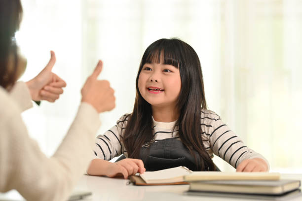 Cute little asian schoolgirl doing homework with happy mother. Distance education, homeschooling concept. stock photo