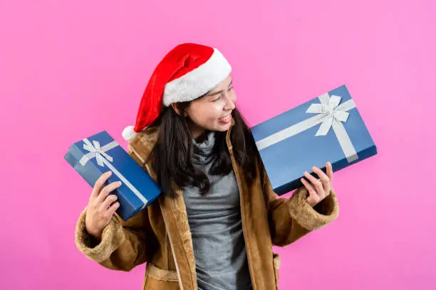 portrait, Asian woman wearing long-sleeved outerwear wearing Christmas hat, holding two gift boxes with both hands use eyes look at box happily with smile, Isolated indoor studio on pinkbackground.
