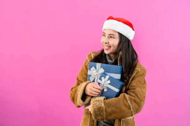 portrait, Asian woman wearing long-sleeved outerwear wearing Christmas hat, wrapped her arms around two gift boxes on chest happily smiling happily, Isolated indoor studio on pinkbackground.