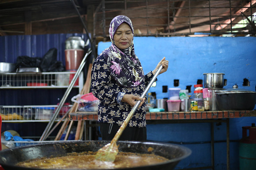 A Malay woman is stirring a big pot of rendang (slow cooked and braised coconut milk with herbs and spices)