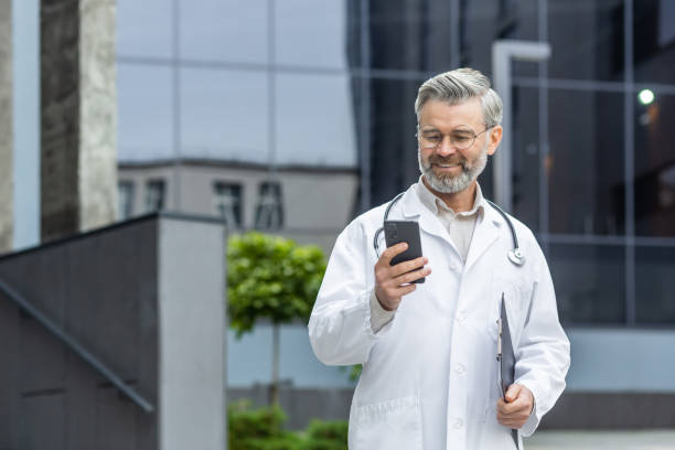 un médico senior con una bata blanca, con un estetoscopio y un paquete en sus manos se para fuera de la clínica y usa el teléfono, escribe un mensaje, habla en una videollamada, sonríe - vertical caucasian glasses red hair fotografías e imágenes de stock